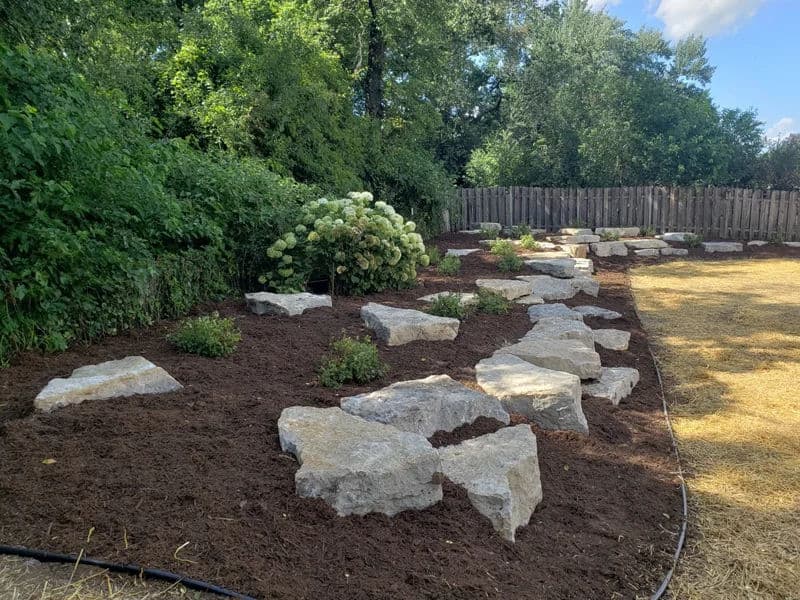 Landscaped yard with large stones, mulch, and flowering plants in a lush green setting.