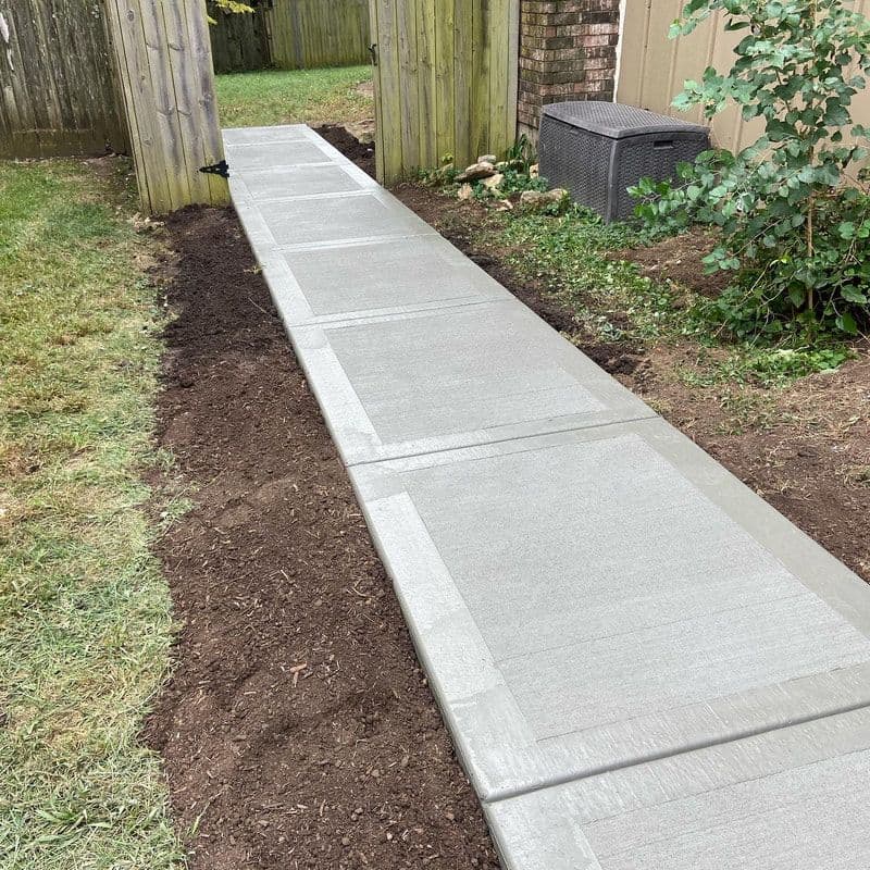 New concrete walkway installation between wooden fences in a landscaped yard.