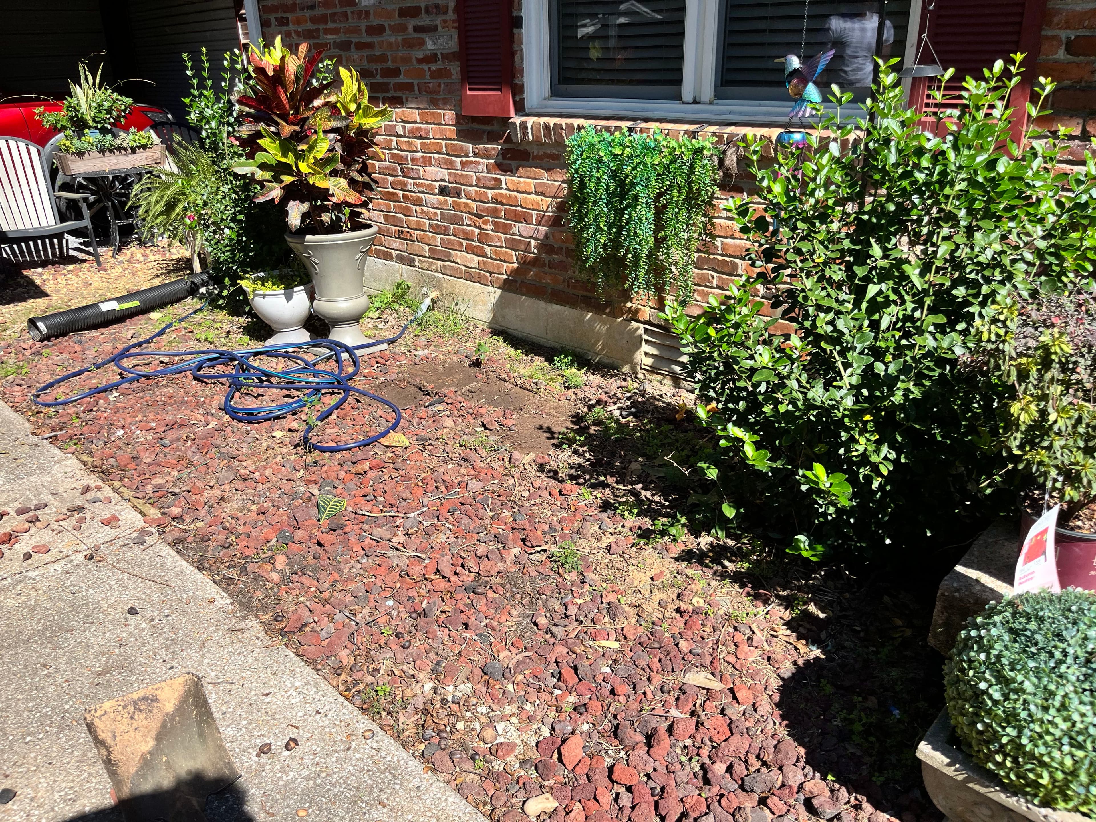 Front yard landscaping featuring red stones, potted plants, and a hose near a brick wall.