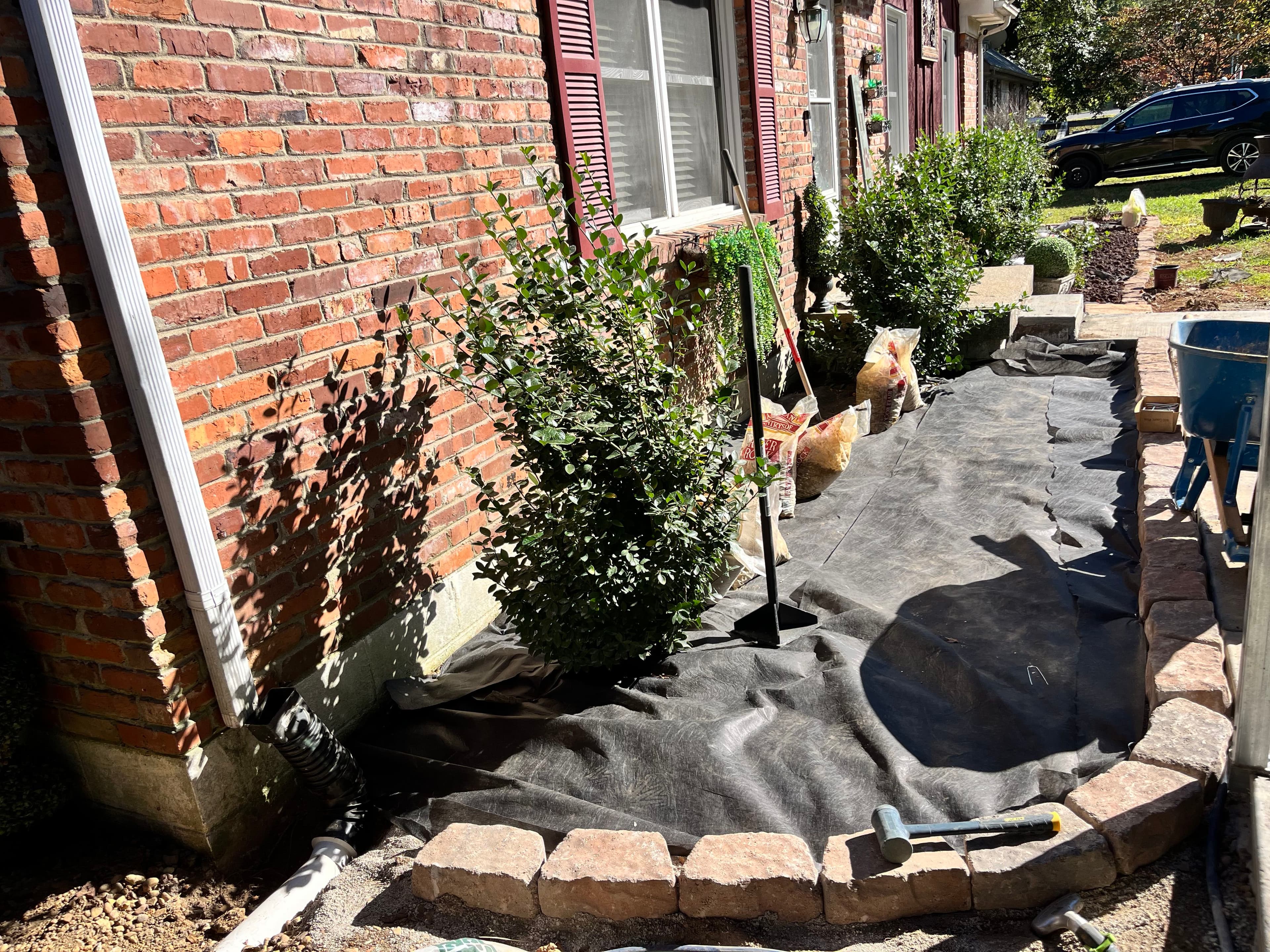 Landscaped garden area with shrubs, garden tools, and a brick border beside a brick house.