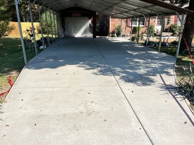 Carport with freshly poured concrete surface, shaded by a roof and surrounded by greenery.