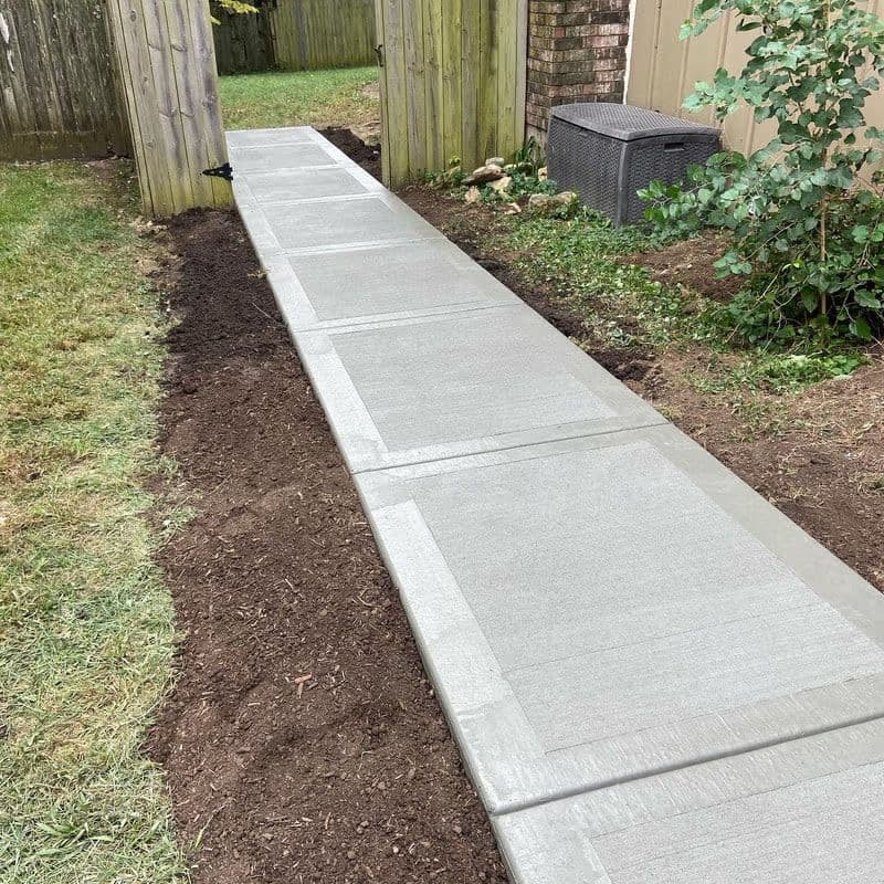 Newly poured concrete walkway in a backyard, surrounded by grass and garden elements.