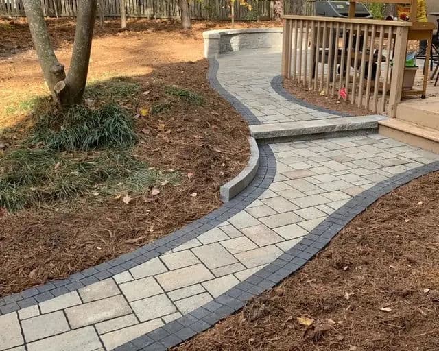 Curved stone walkway with black border leading to a wooden deck in a landscaped yard.