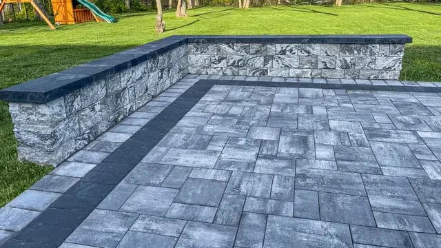 Patio with gray and black stone pavers and a stone wall in a lush green yard.